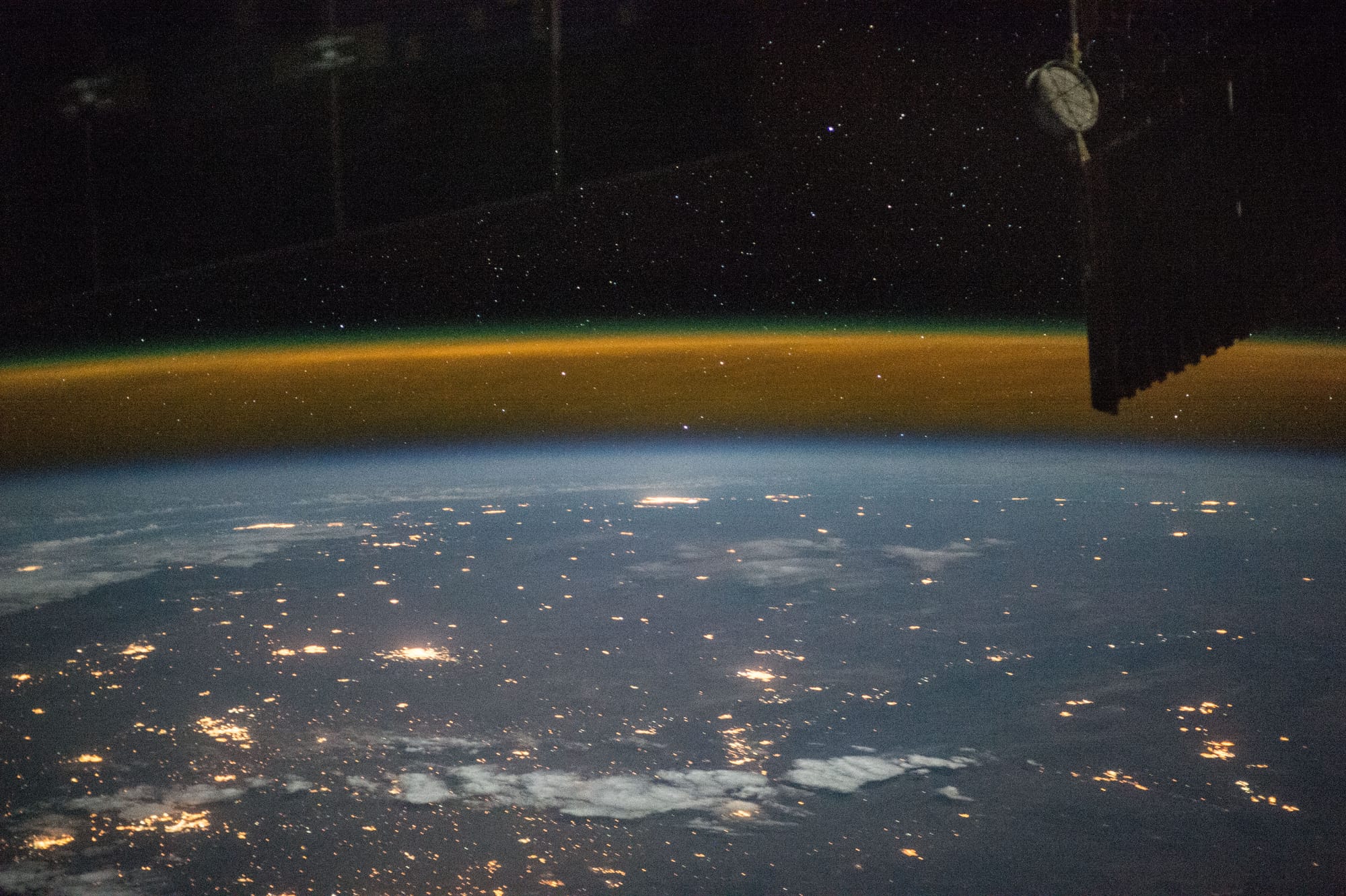 An aerial or space photography of earth from the ISS showing city lights and a big orange line above earth, denoting the Karman Line